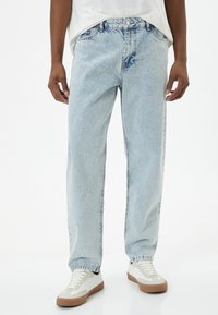 Pantalones vaqueros de mezclilla azul claro con un corte relajado, que presentan parches desgastados, un cierre de botón y un diseño estándar de cinco bolsillos.