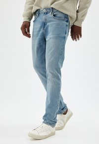 Jeans in denim azzurro chiaro con design a gamba dritta, leggera sfumatura, chiusura con bottone metallico e stile a cinque tasche, abbinati a scarpe da ginnastica bianche.