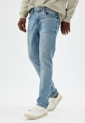 Jeans de denim azul claro con un diseño de pierna recta, ligero desvanecimiento, cierre de botón metálico y estilo de cinco bolsillos, combinados con zapatillas blancas.