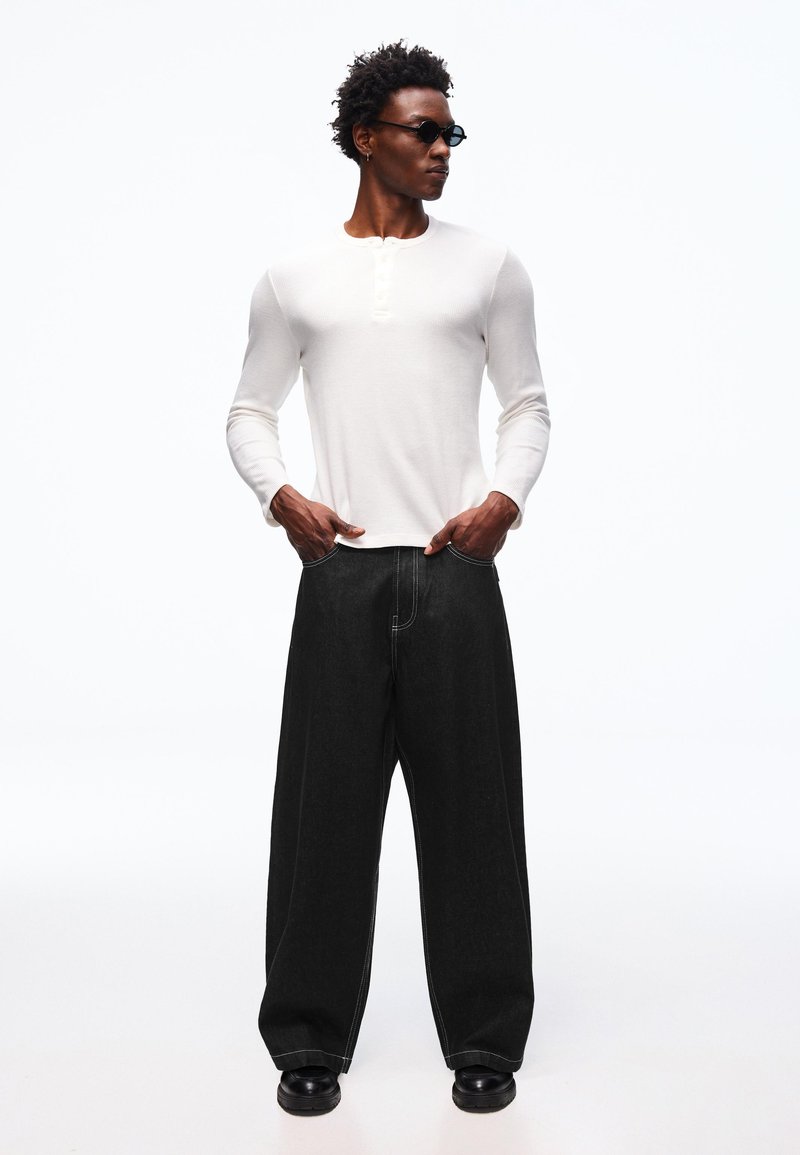 Homme portant des lunettes de soleil noires, une chemise Henley blanche à manches longues, un jean noir à jambes larges et des chaussures noires, debout les mains dans les poches.