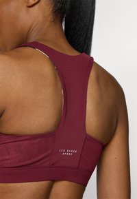 Vīnogu krāsas sporta krūšturis ar sacensību aizmuguri, sieta paneli un smalku logo detaļu; gluda tekstūra ar pieguļošu siluetu.