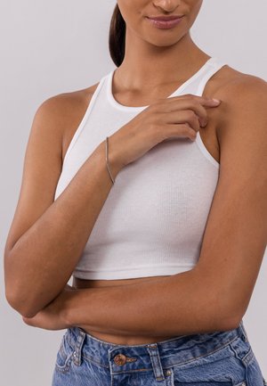 Witte geribbelde crop top en hoge vegan denim shorts. De pols van het model is versierd met een eenvoudige metalen armband; de neutrale achtergrond benadrukt de focus.