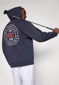 Marineblå hettegenser med et stort sirkulært logo på baksiden som har teksten "USA GOLF," "LOS ANGELES," og "28," med subtil tekstur og kengurulomme.