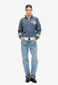 Pull en polaire bleu avec zip à mi-hauteur et lettre "SUPERDRY", manches rayées blanches, porté par-dessus une chemise à col ; associé avec un jean bleu clair.
