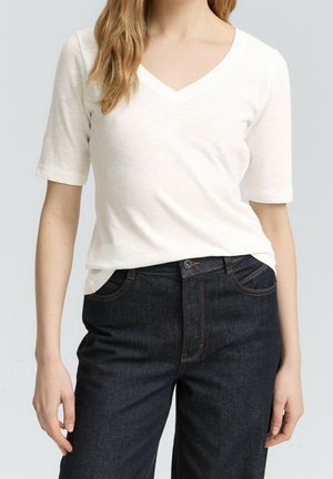 Woman wearing a white V-neck short-sleeve top tucked into high-waisted dark blue jeans against a plain background.