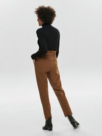 Mujer con cabello rizado vestida con un suéter de cuello alto negro, pantalones marrones de cintura alta y botines negros, de pie con las manos en los bolsillos, de perfil.