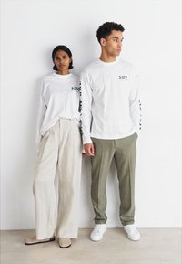 Weiße Langarmhemden mit Text, beige weit geschnittene Hosen und grüne Hosen. Das Schuhwerk umfasst beige Mules und weiße Sneakers. Ein schlichter Hintergrund.