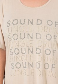 Beige bomulds t-shirt med teksten "SOUND OF JUNGLE DRUM" i metallnitter, skiftende sølv- og guldfarver, med rund halsudskæring.