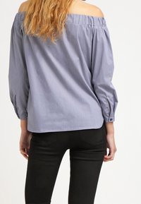 Ljusblå, off-the-shoulder blus med långa ärmar och rynkad halsringning; har knäppningar i ärmsluten och en avslappnad passform.