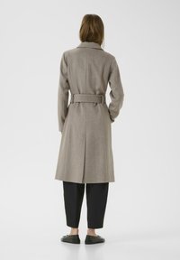 Donna in piedi con la schiena visibile, indossa un cappotto beige con cintura, pantaloni neri a un quarto e scarpe basse nere su uno sfondo bianco semplice.