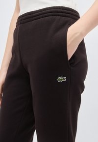 Άτομο που φοράει μαύρο παντελόνι φόρμας με ελαστική μέση και μικρό πράσινο  λογότυπο κροκοδείλου στο μηρό, με το χέρι στην τσέπη.