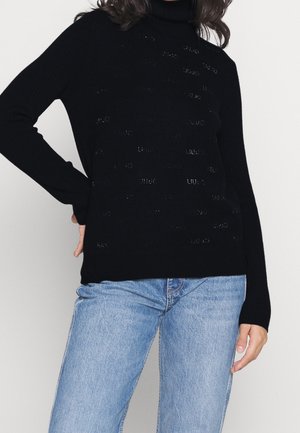 Žena v černém roláku s drobnými perforovanými body a světle modrých džínách, stojící s jednou rukou na boku.