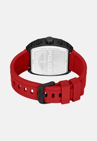 Raudonas silikono laikrodžio dirželis su nerūdijančio plieno korpusu, juodais akcentais ir graviruota nugarėle. Yra kelios reguliavimo skylės ir sagtis.