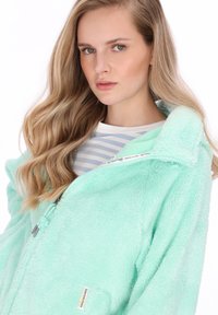 Menta zöld polár dzseki magas gallérral, cipzáros lezárással és puha textúrával, apró címkével. A modellen csíkos ing látható alatta.