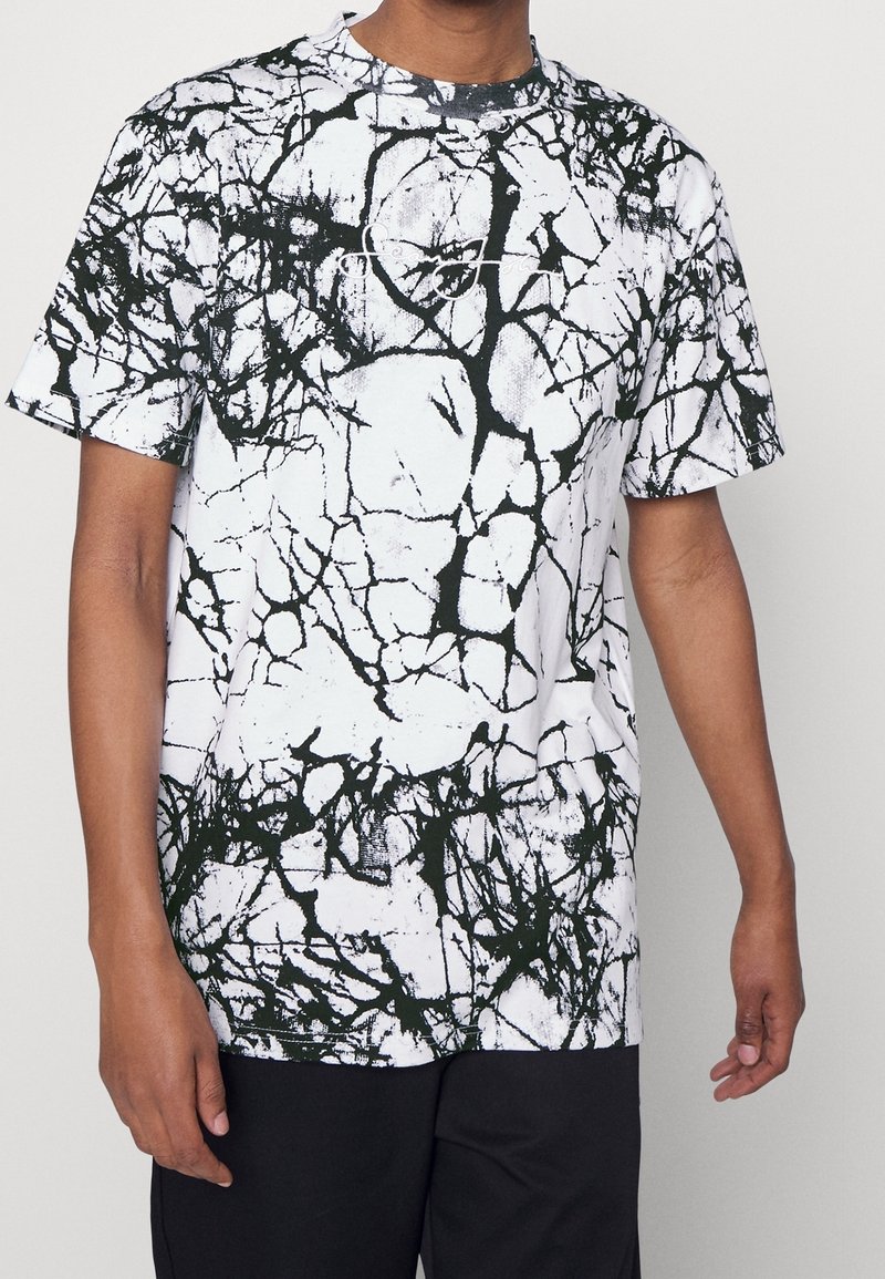 T-shirt blanc à manches courtes avec un motif abstrait noir en effet craquelé. Col rond. Le tissu semble lisse avec une légère texture.