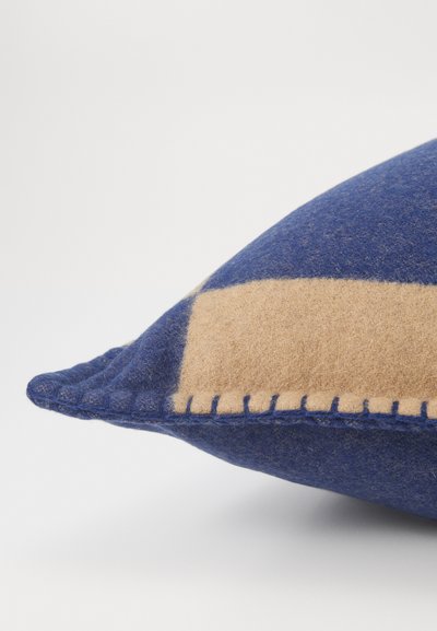 Wollen kussen met een blauwe tint en een tan rechthoekige patch, gedetailleerde stiksels langs de rand en een zachte, textuurafwerking.