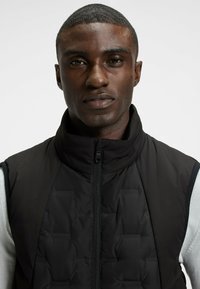 Svart vadderad väst med hög krage, med dragkedja framtill, kontrasterande axlar och subtil märkning. Slät tygtextur, stilren design.