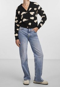 Femme portant un cardigan noir avec des motifs de cœurs beige, un jean droit bleu clair et des chaussures couleur crème, debout avec une main dans la poche.