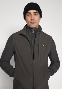 Gilet senza maniche da uomo in grigio scuro, realizzato in tessuto morbido, con chiusura a zip e logo ricamato. Indossato sopra un maglione a coste.