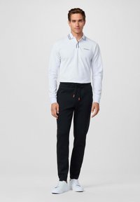 Witte lange mouwen polo met een marineblauw gestreepte kraag, gecombineerd met zwarte joggingsbroeken met een elastische tailleband en een oranje trekkoord. Witte sneakers.