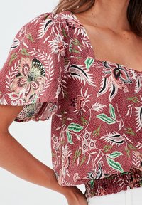 Rote Bluse mit Blumenmuster, rechteckigem Ausschnitt, gepufften Ärmeln und gerafftem Taillenteil. Besitzt grüne und schwarze Akzente aus leichtem Stoff.