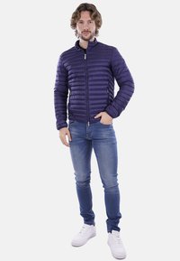 Giacca trapuntata blu navy con zip frontale, colletto rialzato e dettagli di cuciture orizzontali, indossata sopra jeans blu chiaro e sneaker bianche.