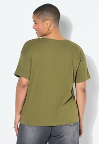 Osoba s krátkými vlasy, oblečená v olivově zeleném tričku a šedých kalhotách, mírně se otočila a usmívá se na pozadí bílé stěny.