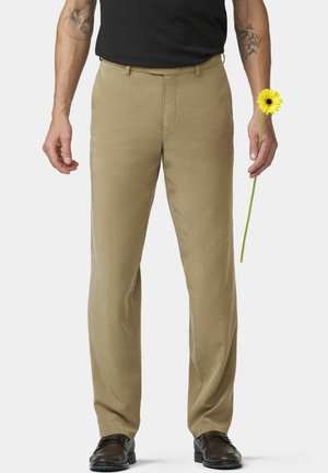 Man met beige broek en zwart shirt houdt een enkele langstengelige gele bloem in zijn rechterhand tegen een effen achtergrond.