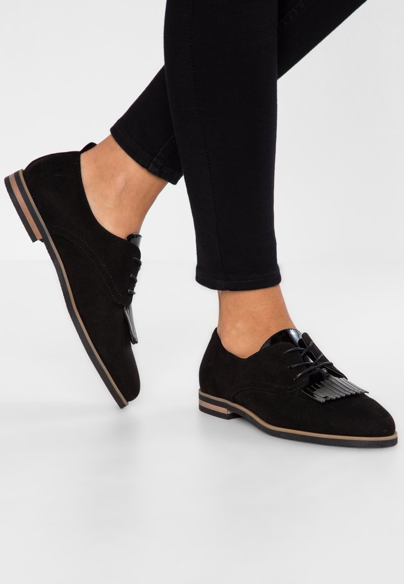 Chaussures à lacets en daim noir avec un accent noir brillant, un détail à franges texturé, et une semelle plate bordée de marron.