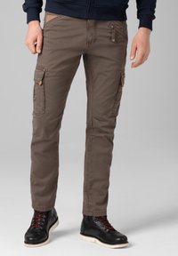 Cargo pants in earthy brown, featuring multiple pockets, a zipper closure, and a slim fit. Paired with black boots, showcasing red laces.