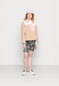 Pullover de fleece em bege, branco e rosa com bolso com fecho; combinado com calções pretos com padrão e ténis brancos; modelo a usar um chapéu de balde branco.