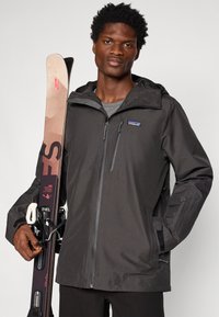 Čierna nepremokavá lyžiarska bunda s kapucňou, predným zipsom a detailmi na vreckách, držaná osobou vedľa lyžiarskej výbavy.