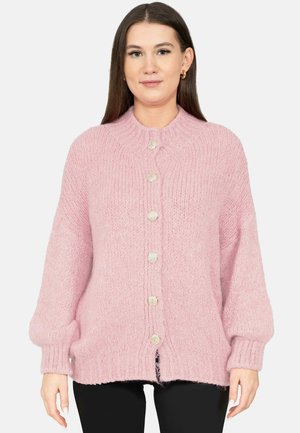 Femme portant un cardigan rose épais boutonné avec des poignets et un col côtelés, associé à un pantalon noir, sur un fond uni.