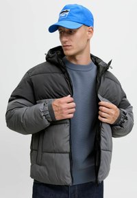 Graue Daunenjacke mit Kapuze, ausgestattet mit elastischen Bündchen und Taschen, über einem blauen Pullover getragen, ergänzt durch eine blaue Mütze.