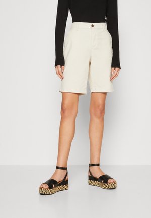 Femme portant un short beige taille haute jusqu'aux genoux, un haut noir à manches longues, et des sandales à plateforme noires avec des semelles tissées.