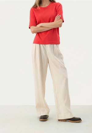 Person trägt ein rotes kurzärmeliges Hemd, weite beige Hosen und dunkelbraune Schuhe, steht mit verschränkten Armen vor einem schlichten Hintergrund.