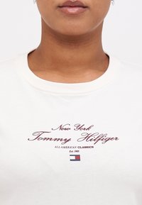 T-shirt en coton blanc avec col rond, présentant un texte imprimé bordeaux : "New York Tommy Hilfiger," et un petit logo coloré en bas.