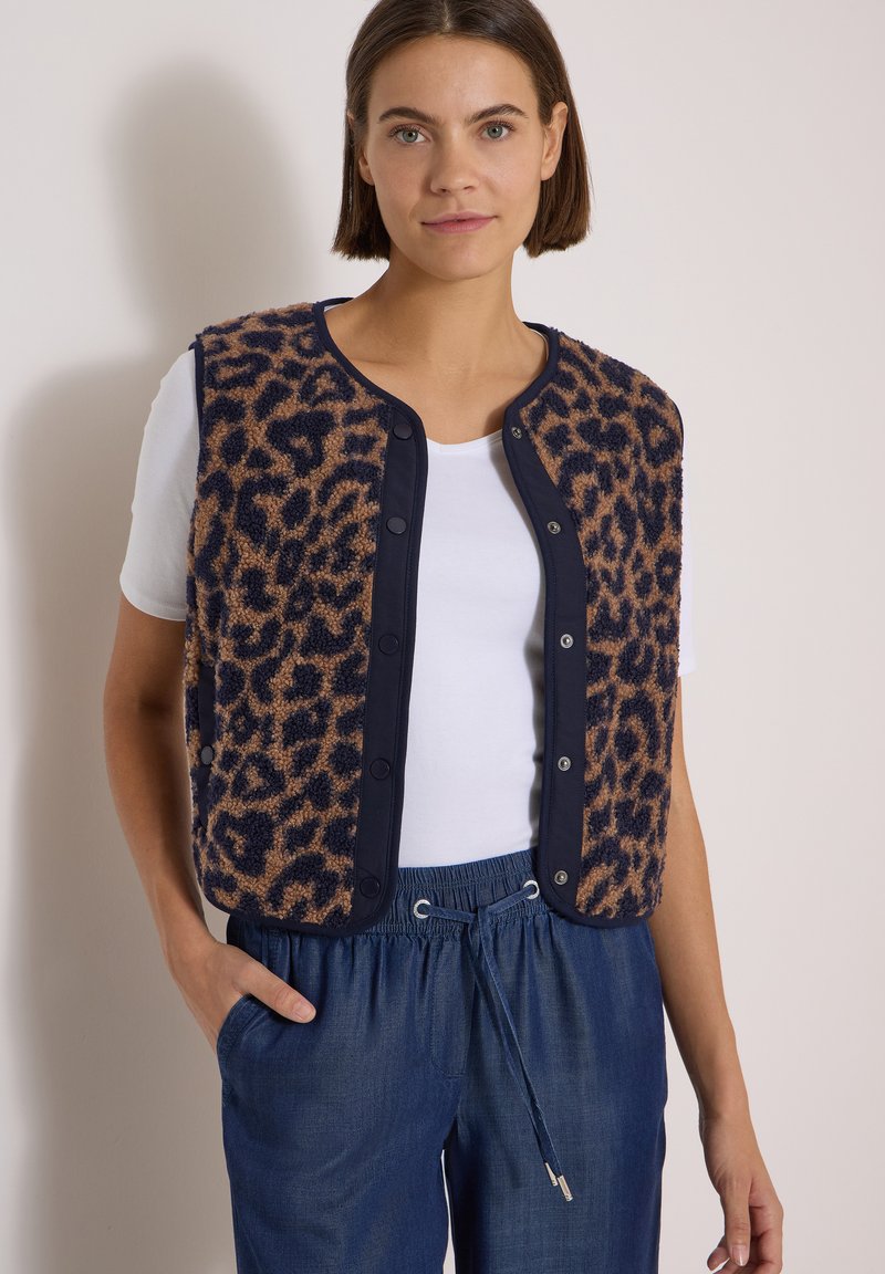 Femme portant un gilet marron et noir à imprimé léopard par-dessus un t-shirt blanc et un pantalon bleu à cordon, debout avec une main dans la poche.