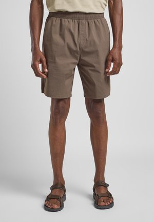 Shorts légers marron avec une taille élastique, des poches latérales et une coupe décontractée, assortis à des sandales marron portées pieds nus.