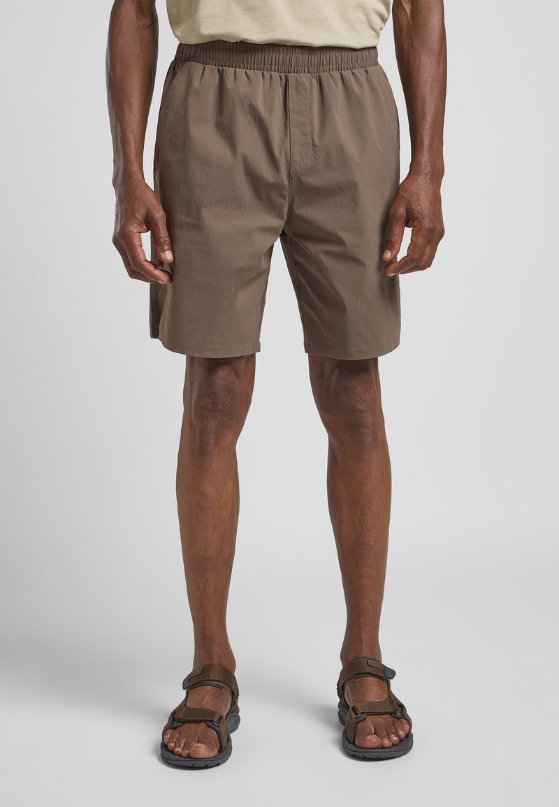 Shorts légers marron avec une taille élastique, des poches latérales et une coupe décontractée, assortis à des sandales marron portées pieds nus.