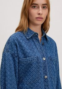 Blaue Jeanshemd mit einer strukturierten Oberfläche, ausgestattet mit einem spitzen Kragen, zwei Brusttaschen und silbernen Knopfverschlüssen.