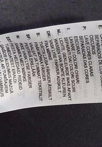 Etichetta di cura per tessuti su tessuto bianco con testo nero, contenente istruzioni in più lingue su come lavare e prendersi cura dei colori.