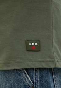 T-shirt verde oliva con un patch rettangolare con "R.D.D." in bianco e un punto rosso. Maniche corte con orlo dritto. Jeans in denim visibili sotto.