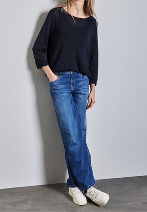 Pull en tricot noir avec un col large, associé à un jean large bleu clair et des baskets blanches. Design simple, sans motifs ni logos visibles.