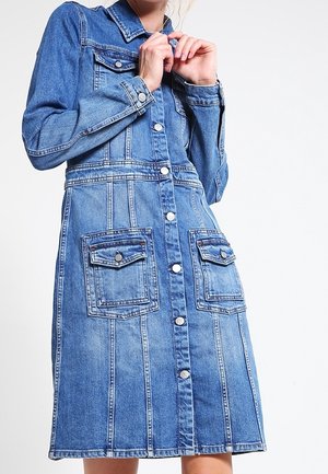 Kvinde iført en mid-lang blå denimkjole med lange ærmer, to knaplommer foran og sølvfarvede knapper foran.
