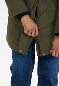 Olivenfarvet pufferjakke med lynlås og knaplukning, synlige ribbede manchetter, båret over blå denimjeans. Fokus på stoftekstur og detaljer.