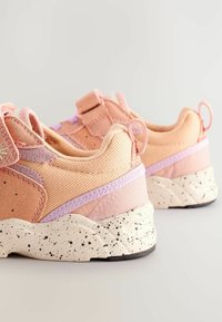 Lyserøde sneakers med en struktureret tekstiloverdel, forstærket hæl og prikket hvid sål. Har justerbare stropper og lilla detaljer.