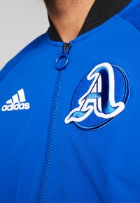 Chaqueta azul con un cuello acanalado negro, que presenta un logotipo bordado en blanco y azul. El detalle de la cremallera es prominente en la parte delantera.