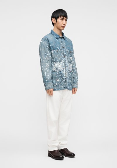 Τζιν μπουφάν με πιτσιλιές από μπογιά, μπλε απόχρωση, τέσσερις μπροστινές τσέπες, κλείσιμο με κουμπιά, συνδυασμένο με λευκό παντελόνι και σκούρα καφέ μπότες.