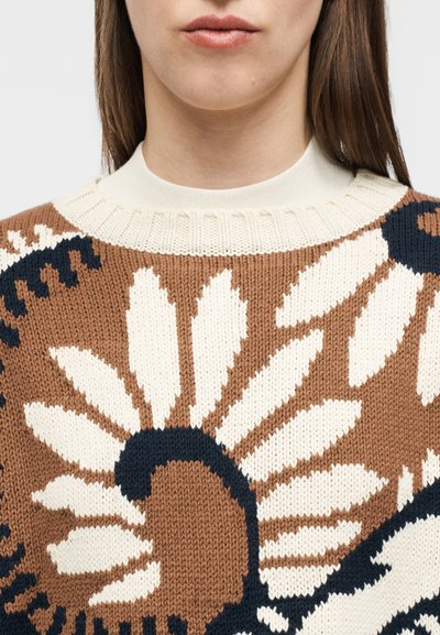 Κοντινή λήψη ενός ατόμου που φοράει καφέ πλεκτό πουλόβερ με μεγάλα λευκά και μαύρα λουλουδάτα σχέδια και εκρού ριμπ γιακά.